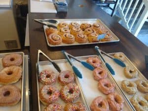 Three trays of various donuts sit on a table with tongs next to each tray.