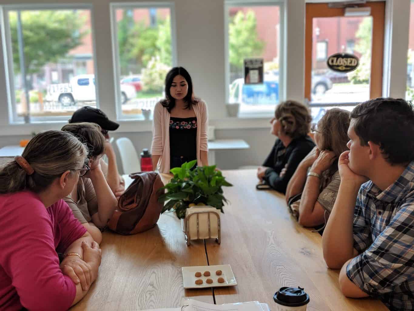 A large group of people sit at a long table listening to a woman talk during an Off the Beaten Path Food Tour in Downtown Lowell.