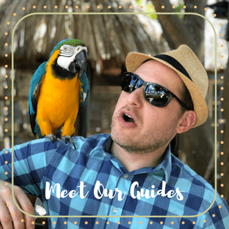 Mark, an Off the Beaten Path Food Tour guide, wears a blue checkered shirt, straw hat, and black sunglasses, while a parrot rests on his shoulder. 