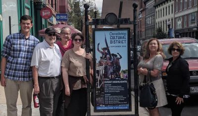 A group of people stand next to a sign in downtown Lowell, MA. 